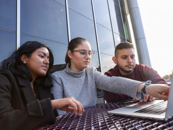 Three students gather around laptop at a picnic table outside and discuss coursework